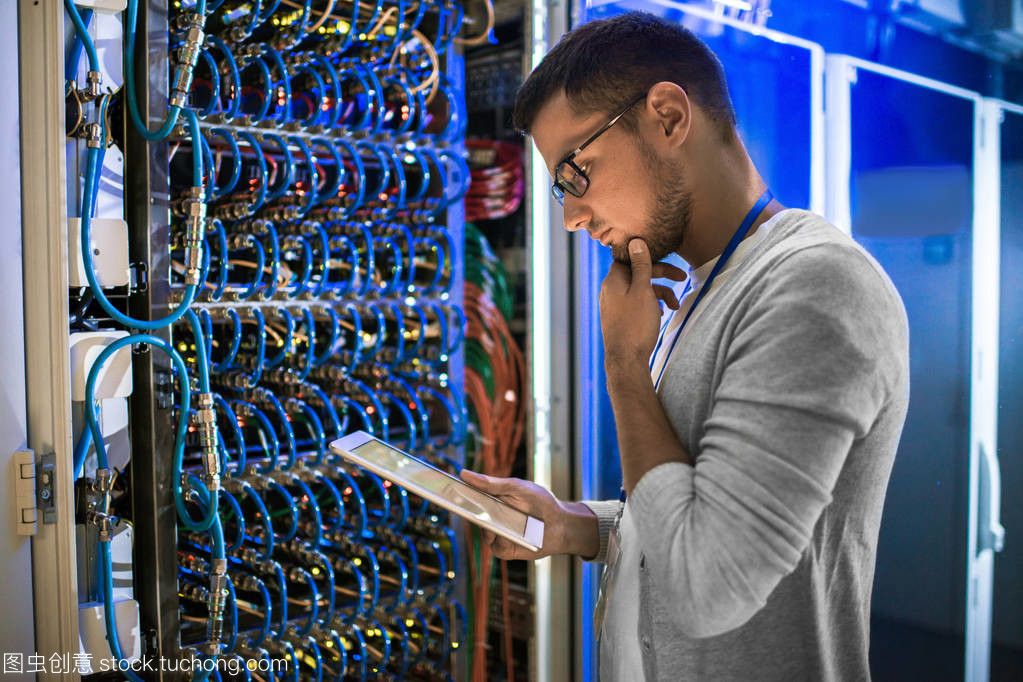 年轻网络工程师的侧面图在数据中心与服务器一起工作时, 若有所思地看着数字平板电脑